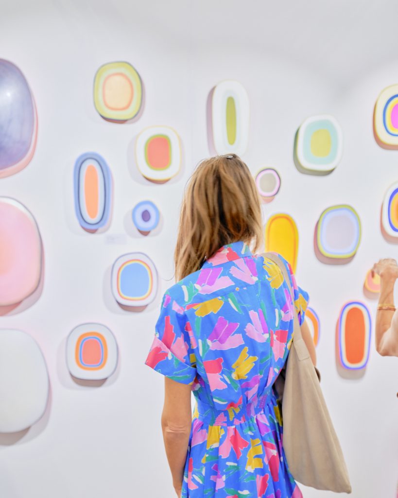 The back of a visitor to the Affordable Art Fair in a colorfully patterned dress looking at a series of small pastel, organically shaped wall mounted artworks.