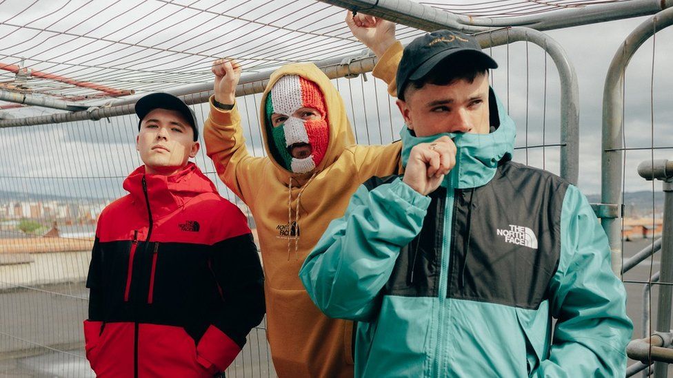 Kneecap made up of three men who use the stage names Mo Chara, Móglaí Bap and DJ Próvaí. The men are all in their 20s. Two wear matching black caps and North Face jackets - one red and the other light blue. The member in the middle wears a yellow hoodie and a knitted balaclava in the colours of the Irish flag. The rappers are pictured outside on a grey day surrounded by fencing, which the middle member holds onto.