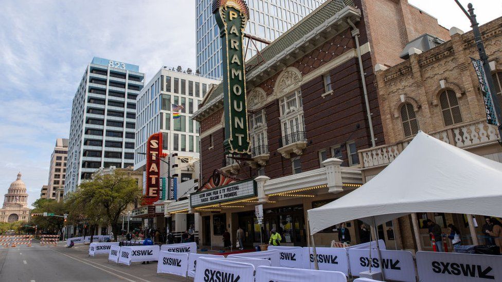 A brick building on a long street with security personnel outside and white barriers in front with SXSW printed on them in black. A large, green vertical sign with the word "Paramount" is attached to the front of the building. In the distance we can see orange and white barriers, showing that the road has been blocked off to allow pedestrians to move around freely.