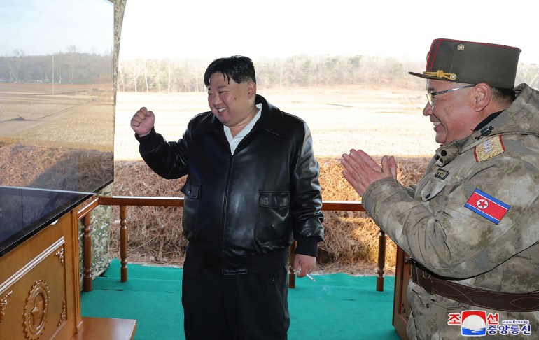 Kim Jong Un punches the air as he watches a screen. A military officer with him appears to be clapping his hands in excitement. Both are smiling.