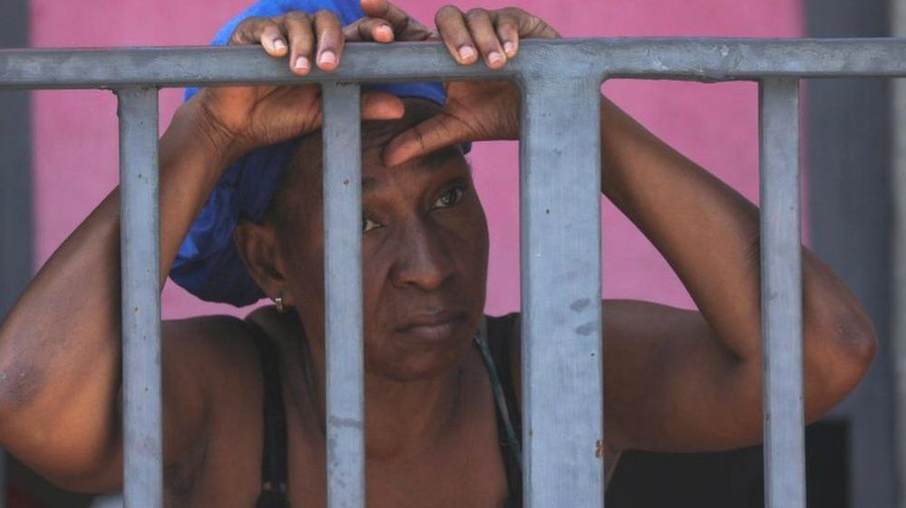 A woman grips a metal fence, a look of sadness on her face