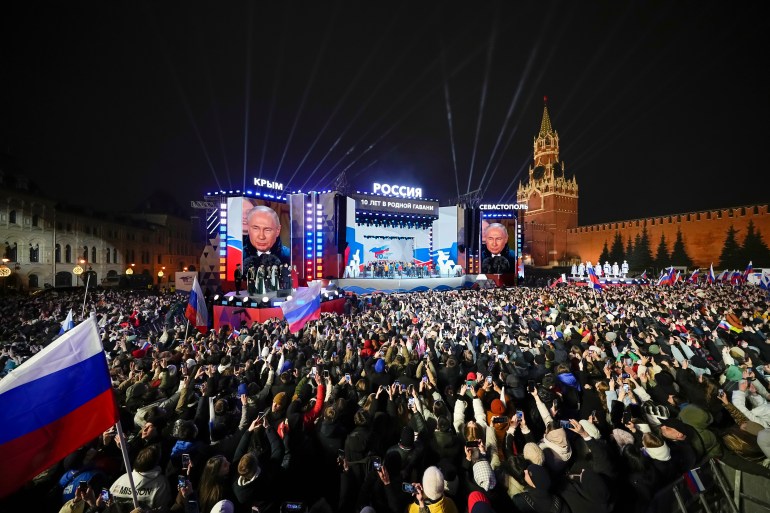 Russian President Vladimir Putin at an event in Red Square. His face is also on massive screens alongside the stage. Some of the crowd are waving Russian flags. The Kremlin is in the background.