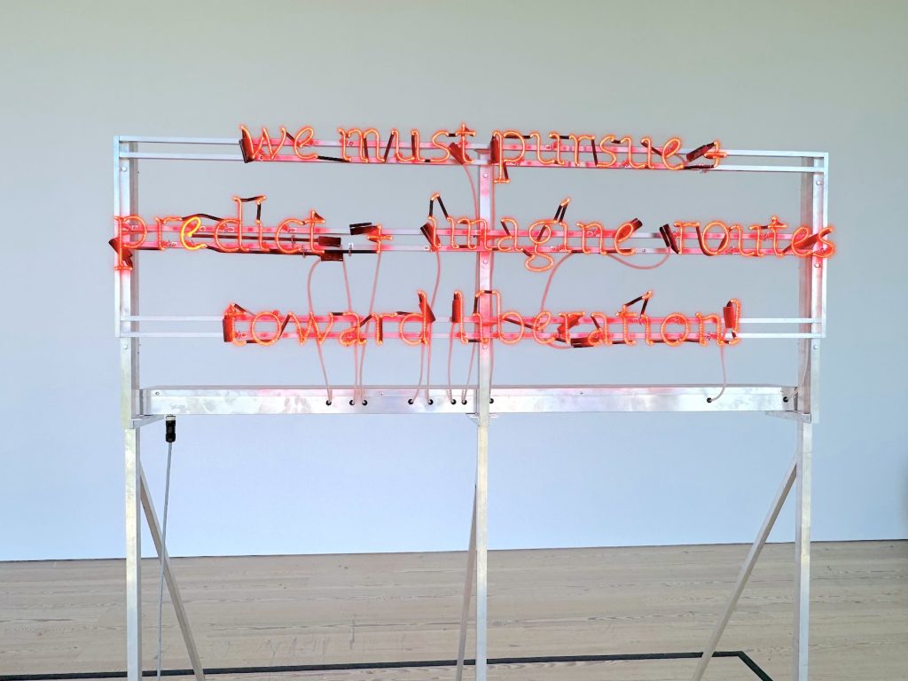 A bright red neon sign in a gallery saying WE MUST PURSUE + PREDICT + IMAGINE ROUTES TOWARD LIBERATION!