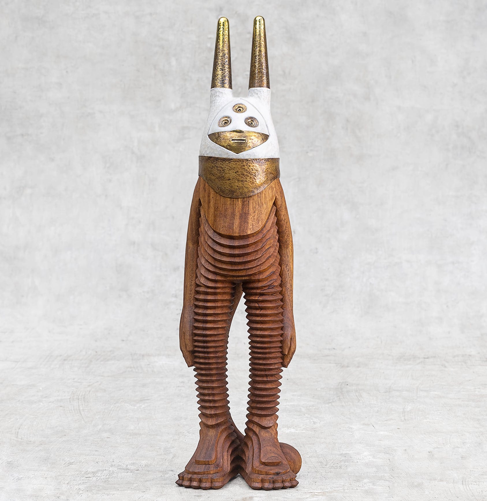 a wooden character with textured legs and a white and gold mask covering its face with three eyes