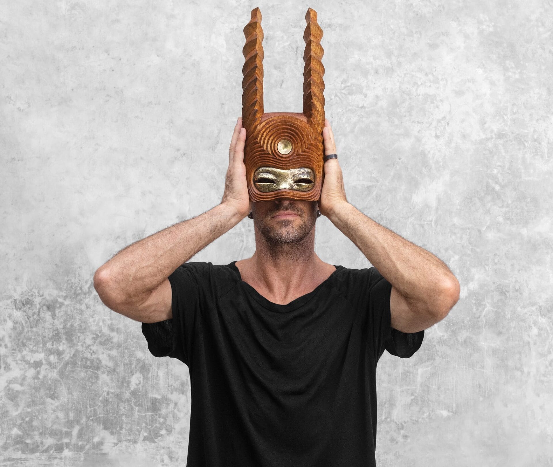 a white man in a black t-shirt stands holding a wooden mask to his face