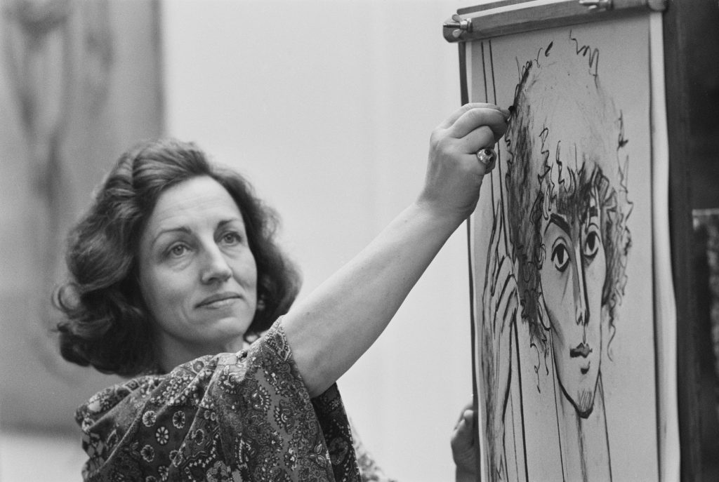 A woman sits at an easel where she sketches a portrait of a woman.